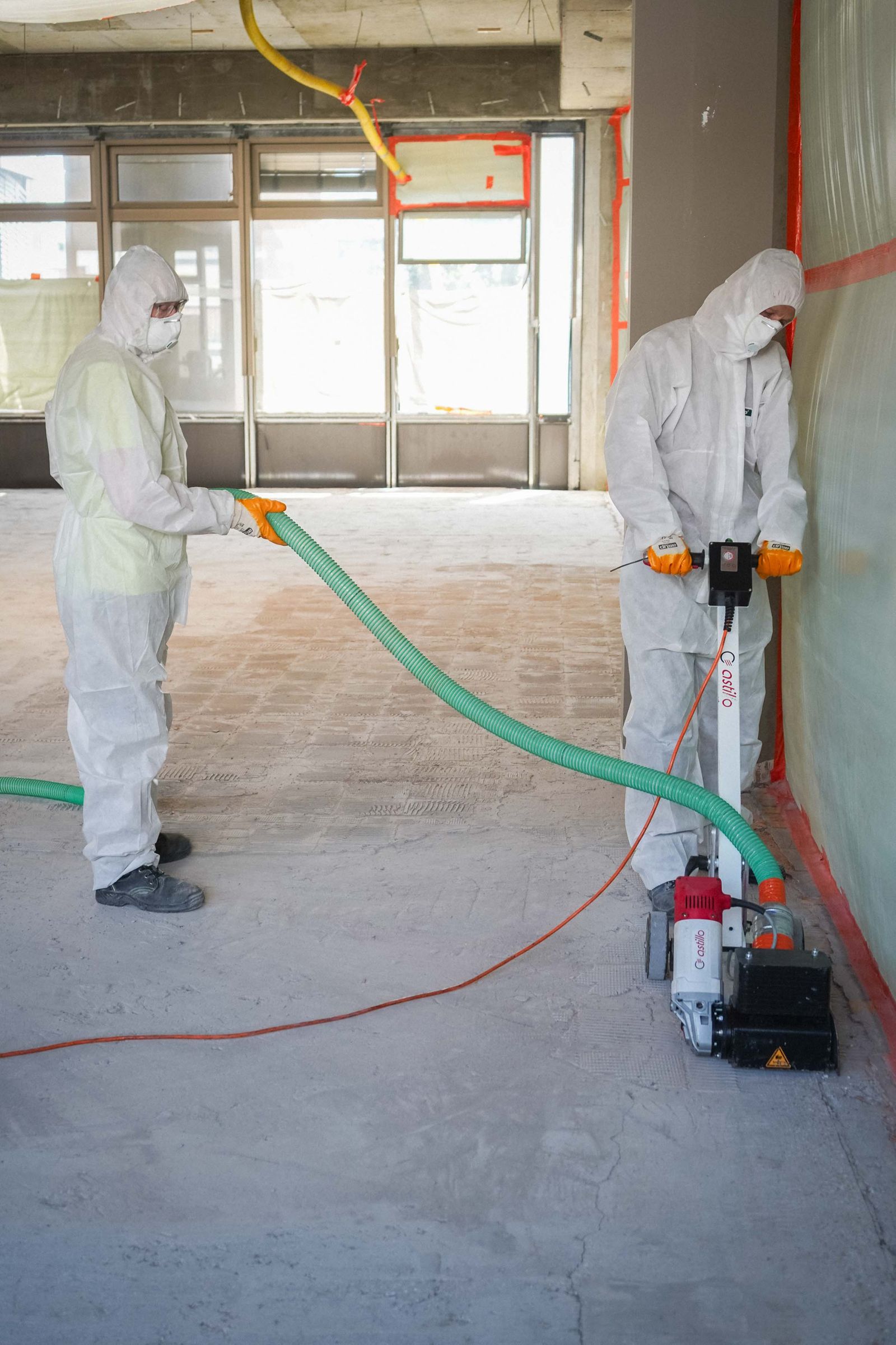 Zwei HSM-Mitarbeiter in weißen Vollschutzanzügen mit Kapuze und Atemschutz bei der Bodensanierung in einem abgeschotteten Asbest-Sanierungsbereich